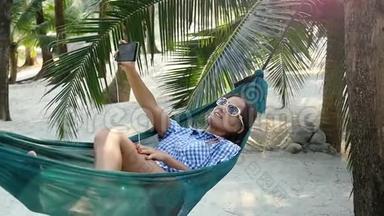 放松的女孩戴太阳镜自拍手机销售手机躺在吊床之间的棕榈树。 慢动作